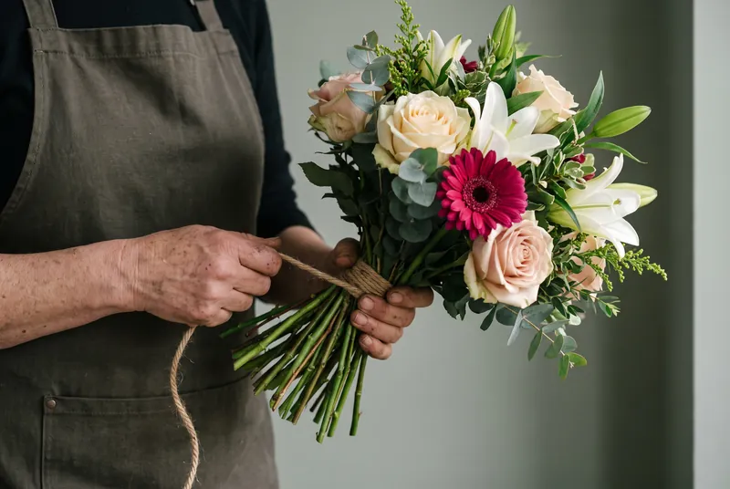 Ein rund gebundener Strauß aus Rosen, Lilien und einer pinkfarbenen Gerbera wird mit Naturgarn gebunden
