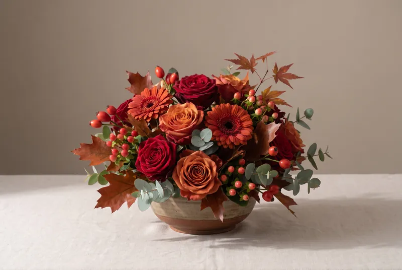 Ein herbstliches Gesteck in dunklem Rot und Orange mit Rosen, Gerbera und Laub in einer flachen Schale